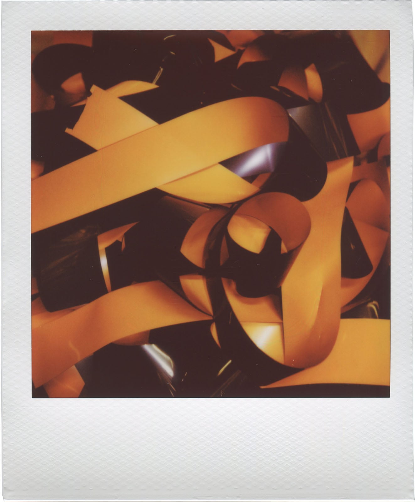 A close-up of tangled orange and black VHS tape ribbons, creating an abstract pattern inside a white Polaroid-style frame.