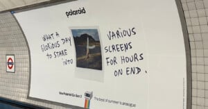 A Polaroid ad in a subway station shows a photo of a person outdoors, with handwritten text saying, "What a glorious day to stare into various screens for hours on end." Polaroid branding and product info are also visible.