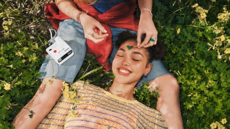 Two people relax on green grass; one lies smiling with eyes closed as the other places a clover leaf on their forehead. A small white camera rests nearby.