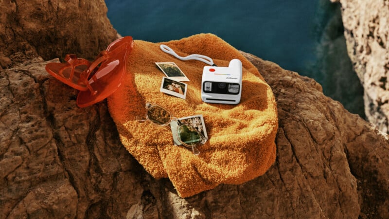 A white Polaroid camera, three instant photos, orange sunglasses, and green-tinted eyeglasses rest on an orange towel spread over rocky cliffs by blue water.