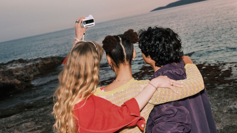 Three friends stand with their arms around each other, facing the ocean at sunset. One person holds up a camera to take a selfie. The rocky shoreline and calm water are visible in the background.