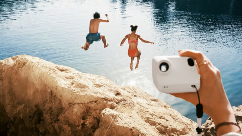 Two people in swimsuits jump into a lake from a rocky ledge while someone in the foreground holds up a portable projector, aiming it toward the jumpers on a sunny day.