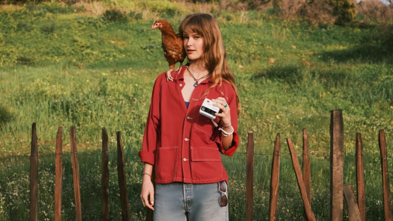 A young woman in a red jacket stands outdoors by a wooden fence, holding a camera. A brown chicken is perched on her shoulder. The background is green with grass and trees.