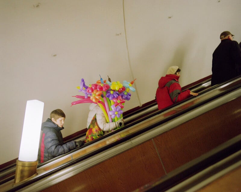 A person on an escalator carries a large bunch of colorful balloon animals. Two other people in winter clothing are riding ahead of them, and a glowing lamp is visible on the left side.