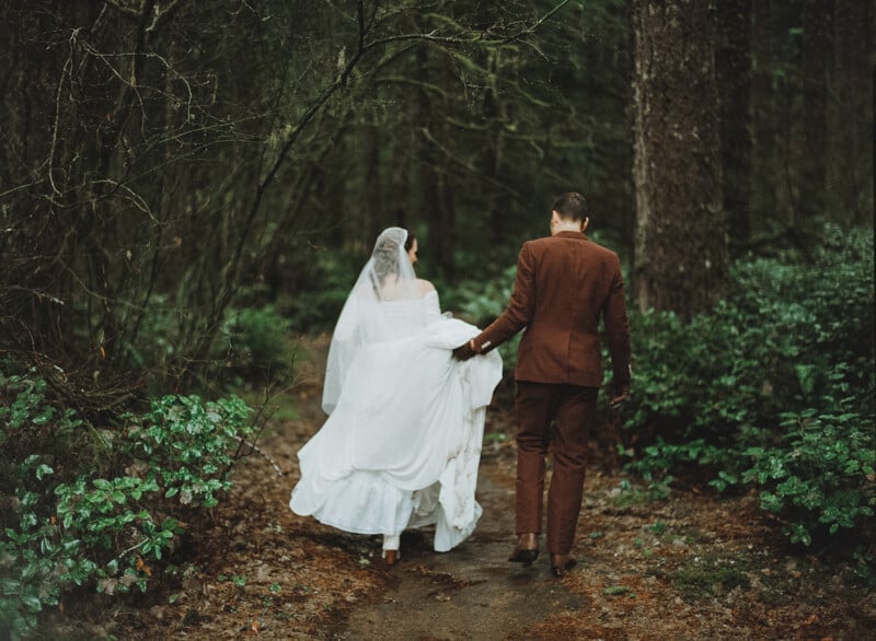 A bride in a white dress and veil walks hand-in-hand with a groom in a brown suit along a forest path, surrounded by green foliage and tall trees.