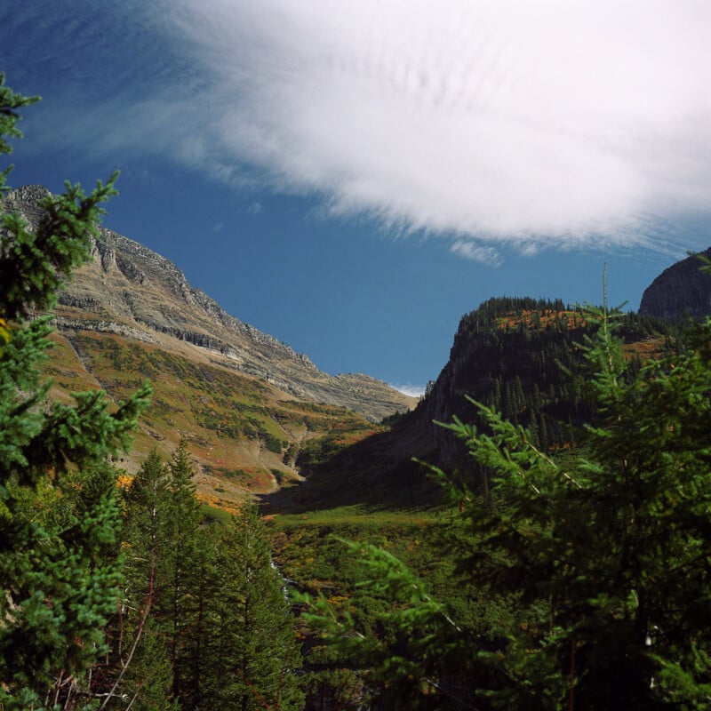 A scenic mountain landscape with green trees in the foreground, sloping hills covered in grass and foliage, and a dramatic cloud stretching across a bright blue sky.