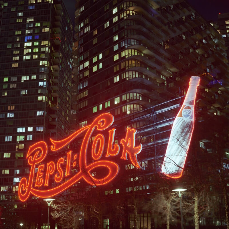 Bright neon Pepsi-Cola sign featuring a bottle glows at night in front of a modern high-rise apartment building with many lit windows.