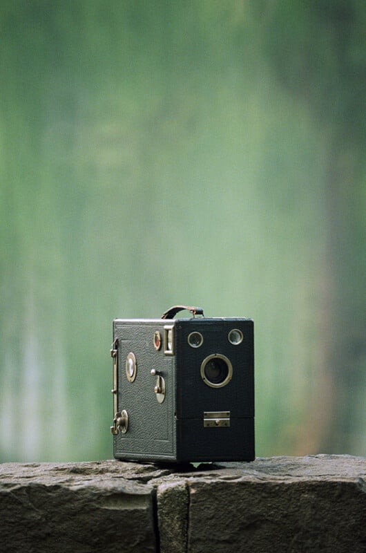 A vintage box camera sits on a stone ledge outdoors, with a blurred green background of trees or foliage.