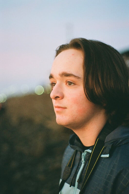 A young person with shoulder-length brown hair looks thoughtfully into the distance while standing outdoors at dusk, wearing a dark jacket. Blurred lights are visible in the background.
