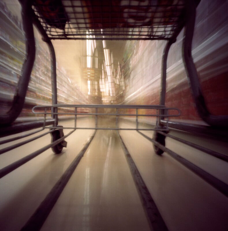 Blurred view from inside a shopping cart looking down a grocery store aisle, with shelves and lights creating a sense of motion and speed.