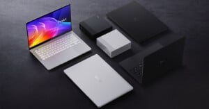 A group of laptops and desktop mini PCs in black and silver colors are arranged on a dark surface; one laptop screen displays a colorful abstract wallpaper.