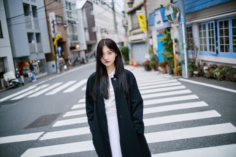 A woman with long black hair wearing a black coat stands on a crosswalk in an urban street, with buildings, shops, and plants visible in the background.