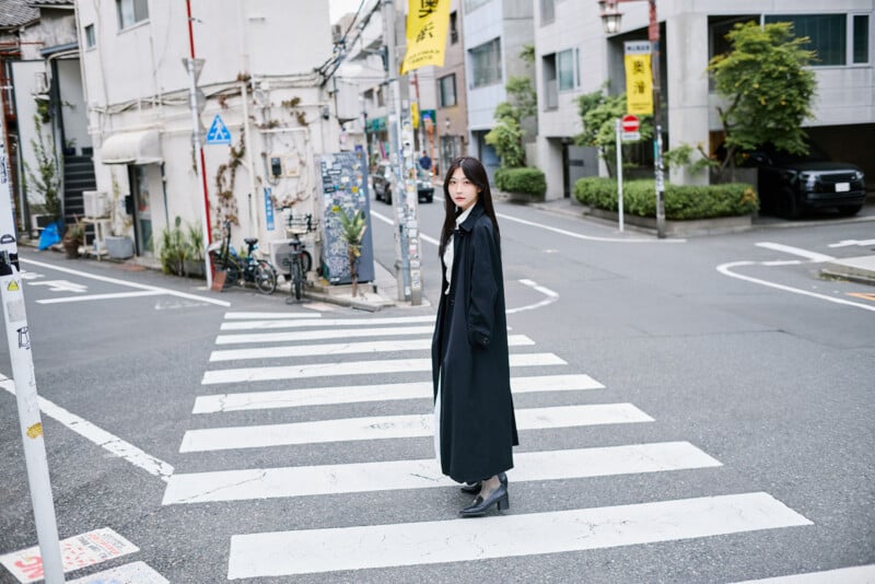 A woman in a long black coat and boots stands on a crosswalk in a quiet urban street, surrounded by buildings, plants, and traffic signs.