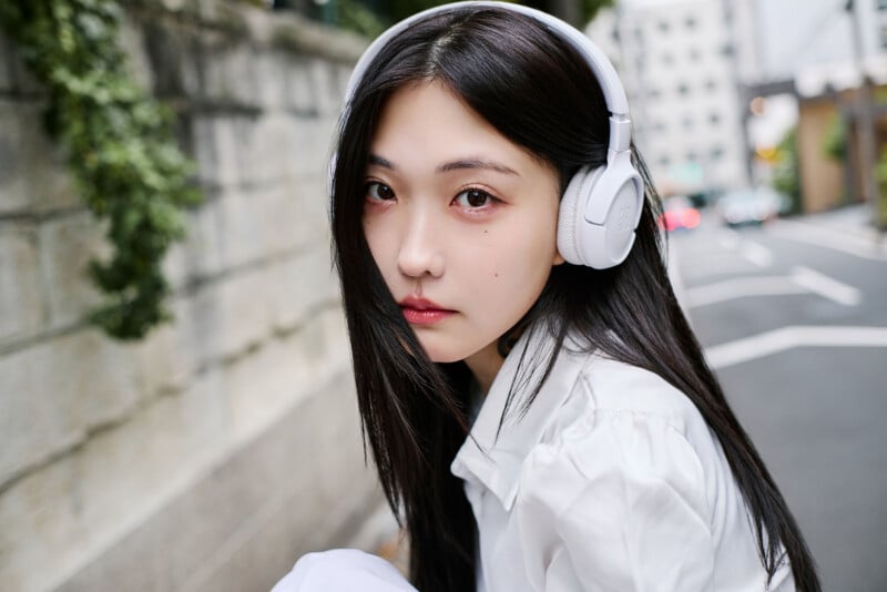 A young woman with long dark hair wearing white headphones and a white shirt looks at the camera while sitting on a street with a stone wall and blurred buildings in the background.
