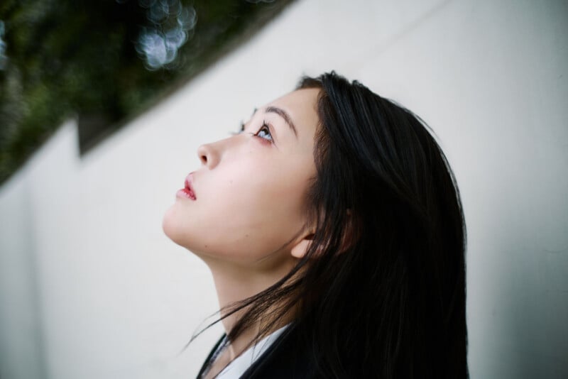 A young woman with long dark hair looks upward thoughtfully while standing against a white wall, with soft natural light illuminating her face.