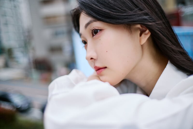 A young woman with long dark hair gazes thoughtfully into the distance, resting her chin on her arms while wearing a white shirt. The background shows an out-of-focus city street and buildings.