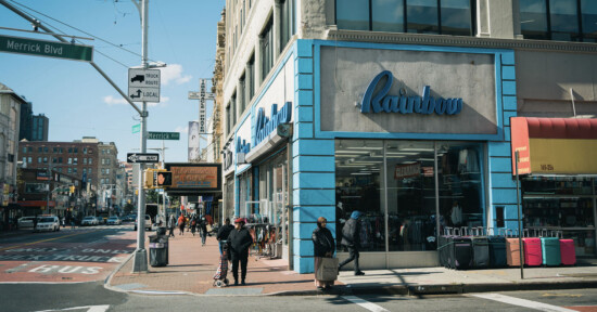 A city street corner with people walking past a store called “Rainbow” that has a blue and white exterior. Street signs for Merrick Blvd and 165th St are visible, along with other shops and a row of luggage outside.