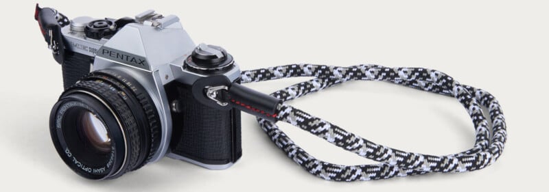A vintage Pentax film camera with a black and silver body is attached to a black-and-white patterned neck strap, displayed on a light background.