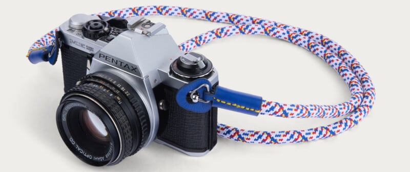 A vintage Pentax film camera with a zoom lens is attached to a colorful, braided camera strap with blue, white, red, and yellow patterns. The camera is positioned on a white background.