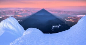 Snowy mountain peaks frame a dramatic triangular shadow cast over a valley at sunrise, with pink and purple tones illuminating the distant landscape and sky.
