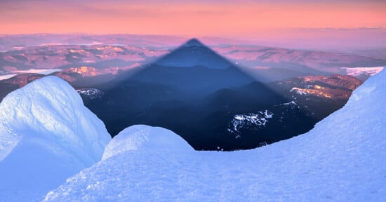 Snowy mountain peaks frame a dramatic triangular shadow cast over a valley at sunrise, with pink and purple tones illuminating the distant landscape and sky.