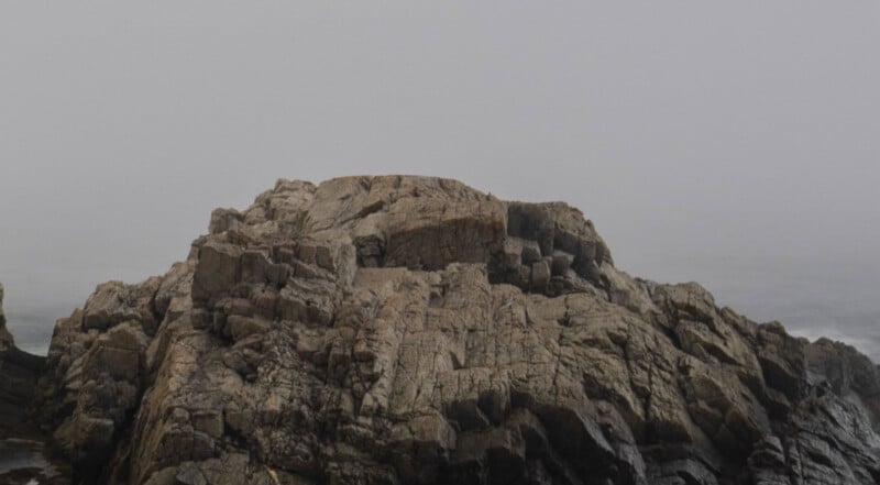 A large, rugged rock formation sits by the ocean under a gray, foggy sky, with faint waves visible in the background.