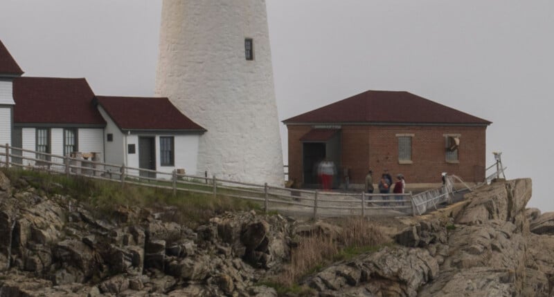 A lighthouse with a white tower and nearby brick and white buildings sits on rocky terrain, with several people standing near the entrance on a cloudy day.