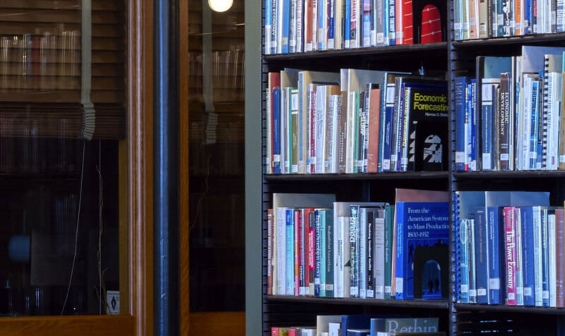 A bookshelf filled with various books stands next to a glass door with wooden frames, reflecting indoor lights. Some book titles are visible, and the setting suggests a quiet library or study area.