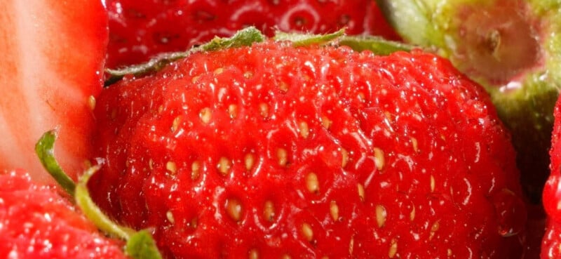 Close-up of a ripe, red strawberry showing its textured surface, small yellow seeds, and green leafy top, with sections of other strawberries visible around the edges.