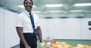 A man in a white shirt, dark tie, and badge smiles while standing beside a table with trays of food and snacks in a brightly lit, modern room with white walls and a green floor.