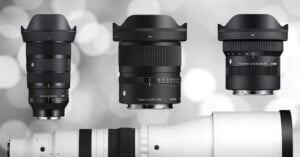 Three black camera lenses with lens hoods are displayed upright against a blurred gray background, next to a large white telephoto lens positioned horizontally at the bottom of the image.