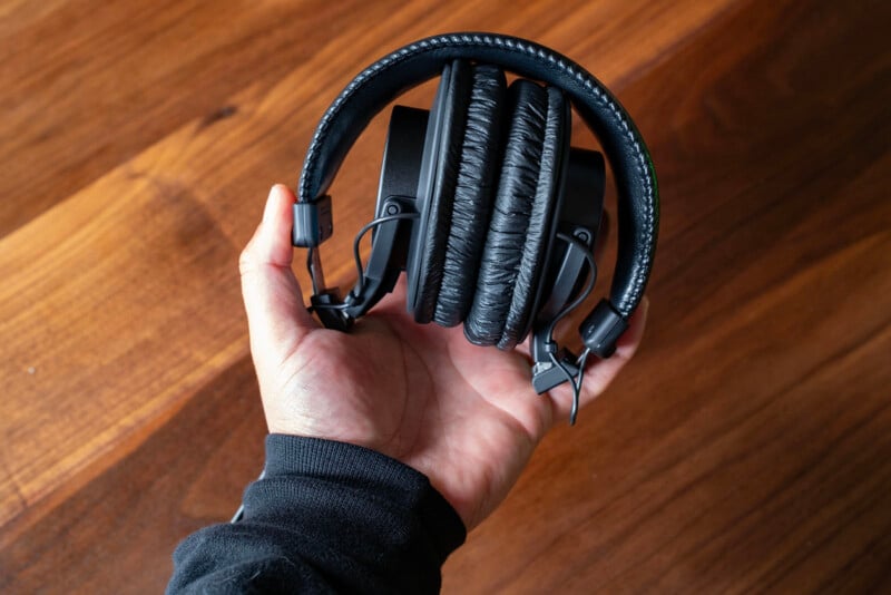 A person wearing a black sleeve holds a pair of black, foldable over-ear headphones with cushioned ear pads over a wooden surface.