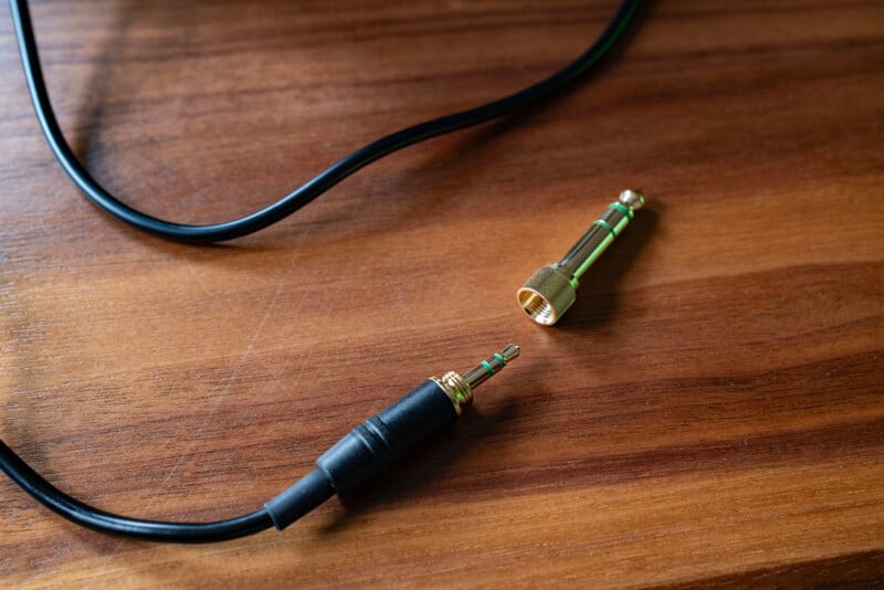 A black audio cable with a 3.5mm jack and a separate gold 6.35mm adapter are lying on a wooden surface.