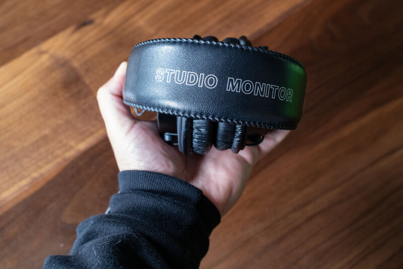 A hand holding a pair of black headphones with "STUDIO MONITOR" printed in white text on the headband, against a wooden background.