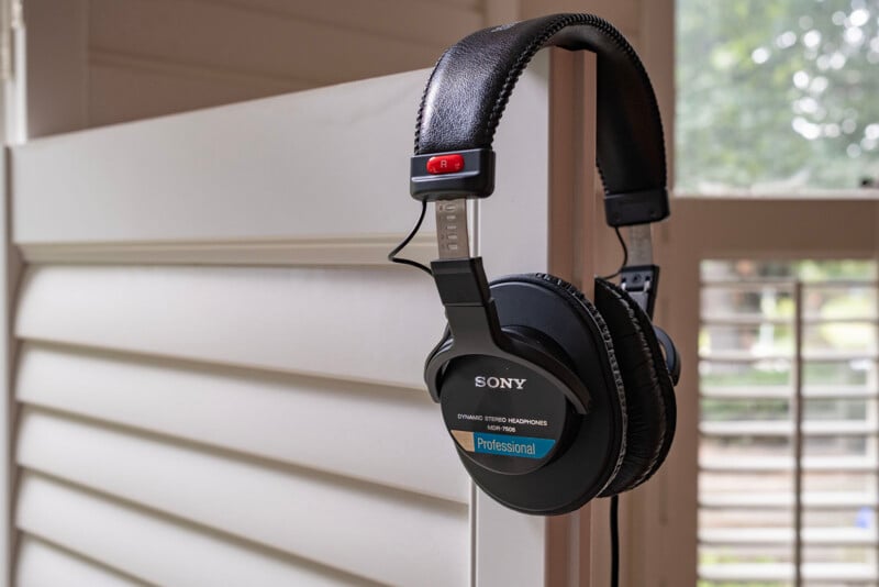 A pair of black Sony professional headphones hangs on the edge of a white shuttered door, with sunlight and green trees visible through a window in the background.