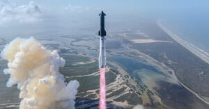 A rocket launches into the sky above a coastal landscape, leaving a trail of smoke and flames behind as it ascends over water and marshland.