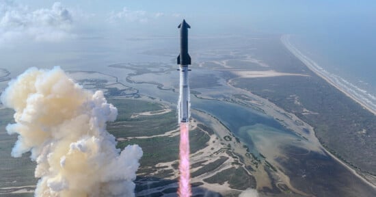A rocket launches into the sky above a coastal landscape, leaving a trail of smoke and flames behind as it ascends over water and marshland.