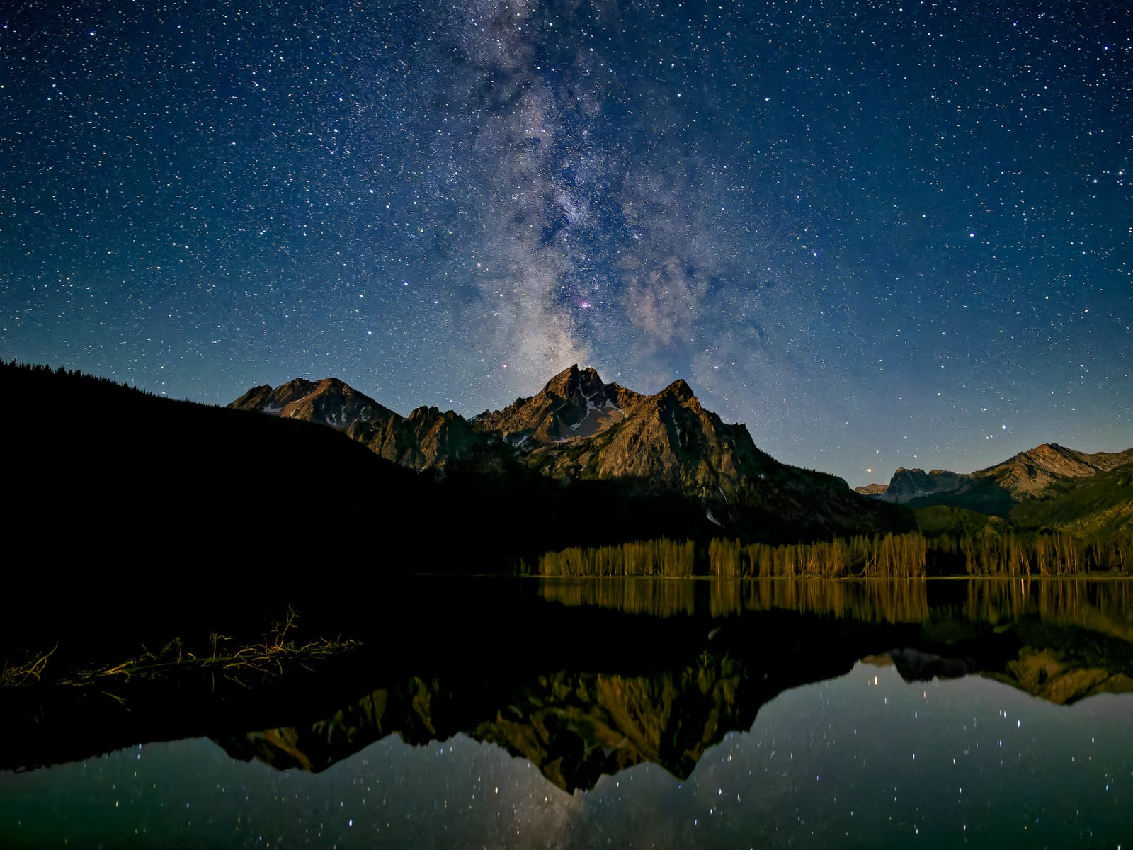 A star-filled night sky with the Milky Way visible above mountain peaks, reflected in a calm lake below. Dense trees line the shore, silhouetted against the horizon.