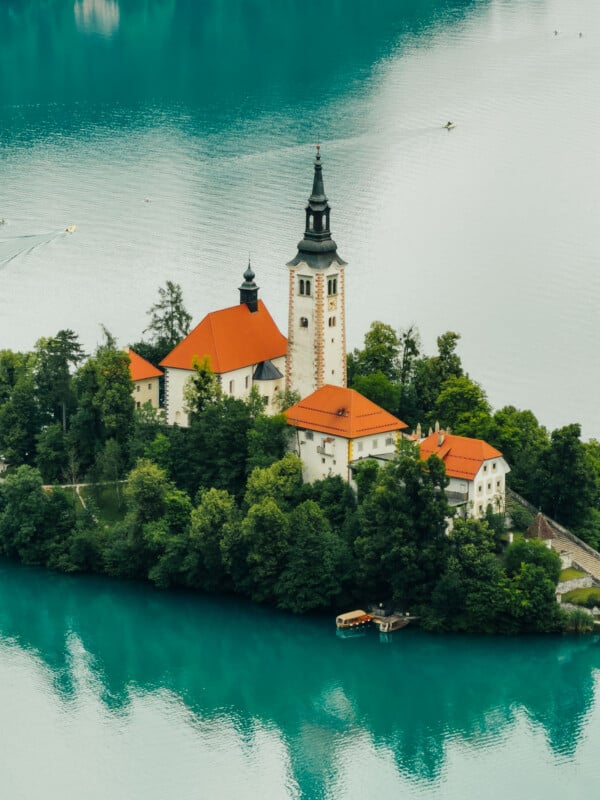 A small island with a church featuring a tall bell tower and red-tiled roofs sits in the middle of a turquoise lake, surrounded by lush green trees and calm water.