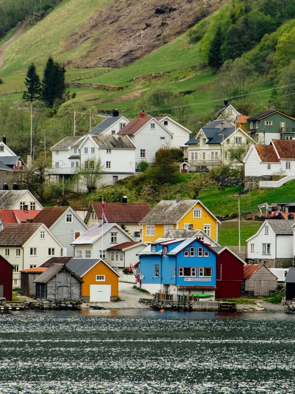Colorful wooden houses, including yellow, blue, red, and white, are clustered along a calm waterfront with a grassy hillside and trees in the background. The scene appears cozy and picturesque.