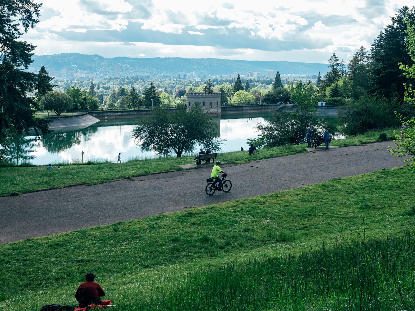 A person rides a bike on a paved path through a green park beside a large reflective reservoir, with people relaxing nearby and a cityscape visible in the background under a partly cloudy sky.