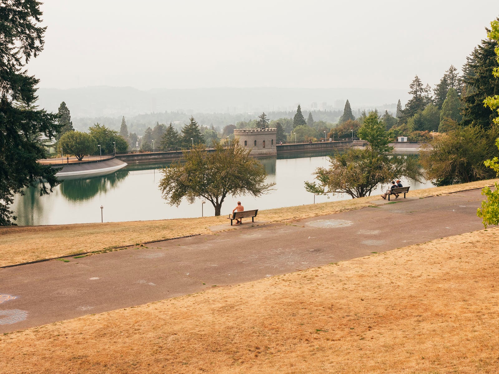 A few people sit on benches facing a calm reservoir surrounded by trees and grass, with a historic stone building in the background and distant hills visible through a hazy sky.