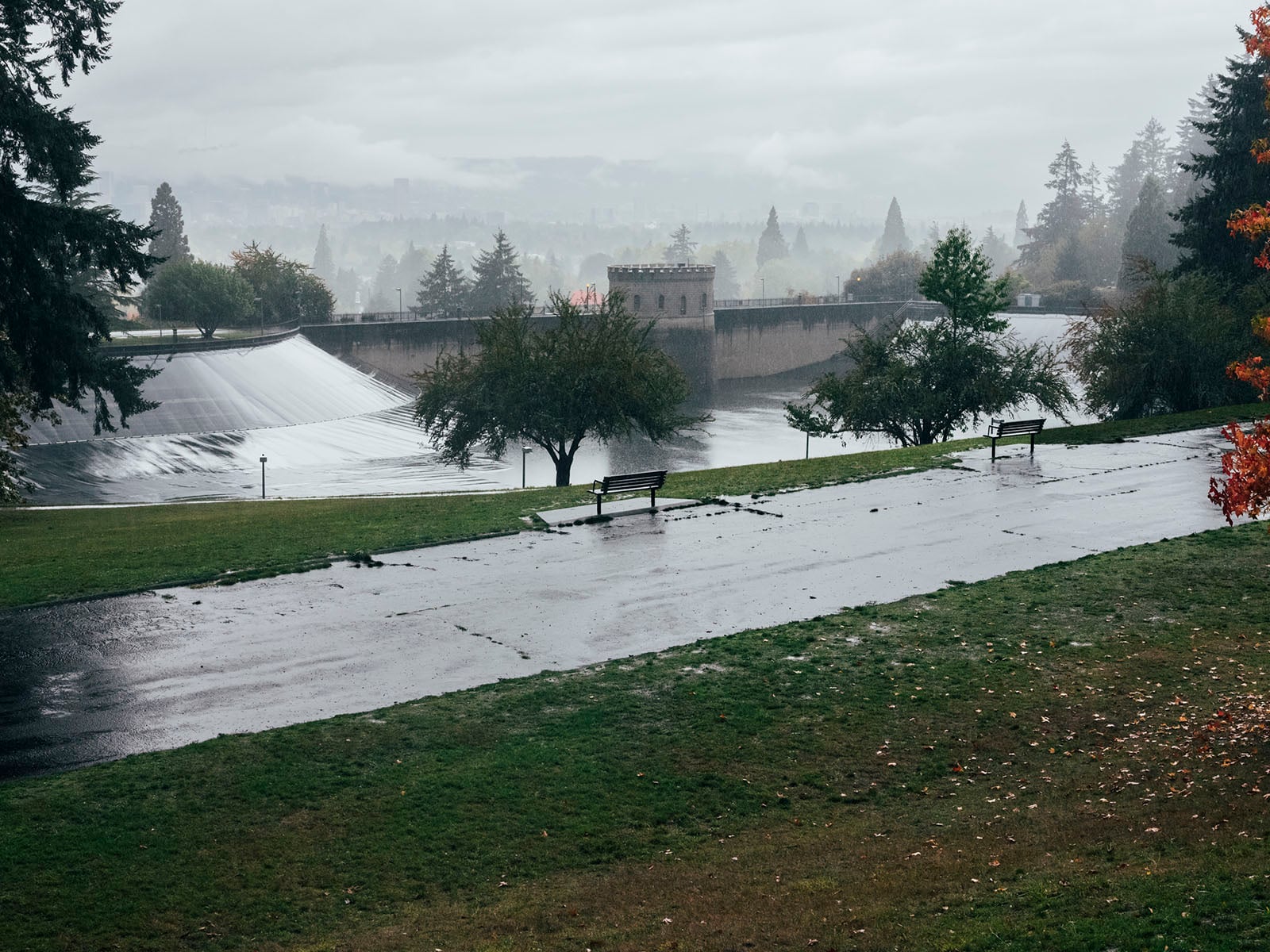 A rainy, overcast day at a park featuring wet pavement, grassy areas, empty benches, trees, and a large dam or spillway in the background with misty hills and trees in the distance.