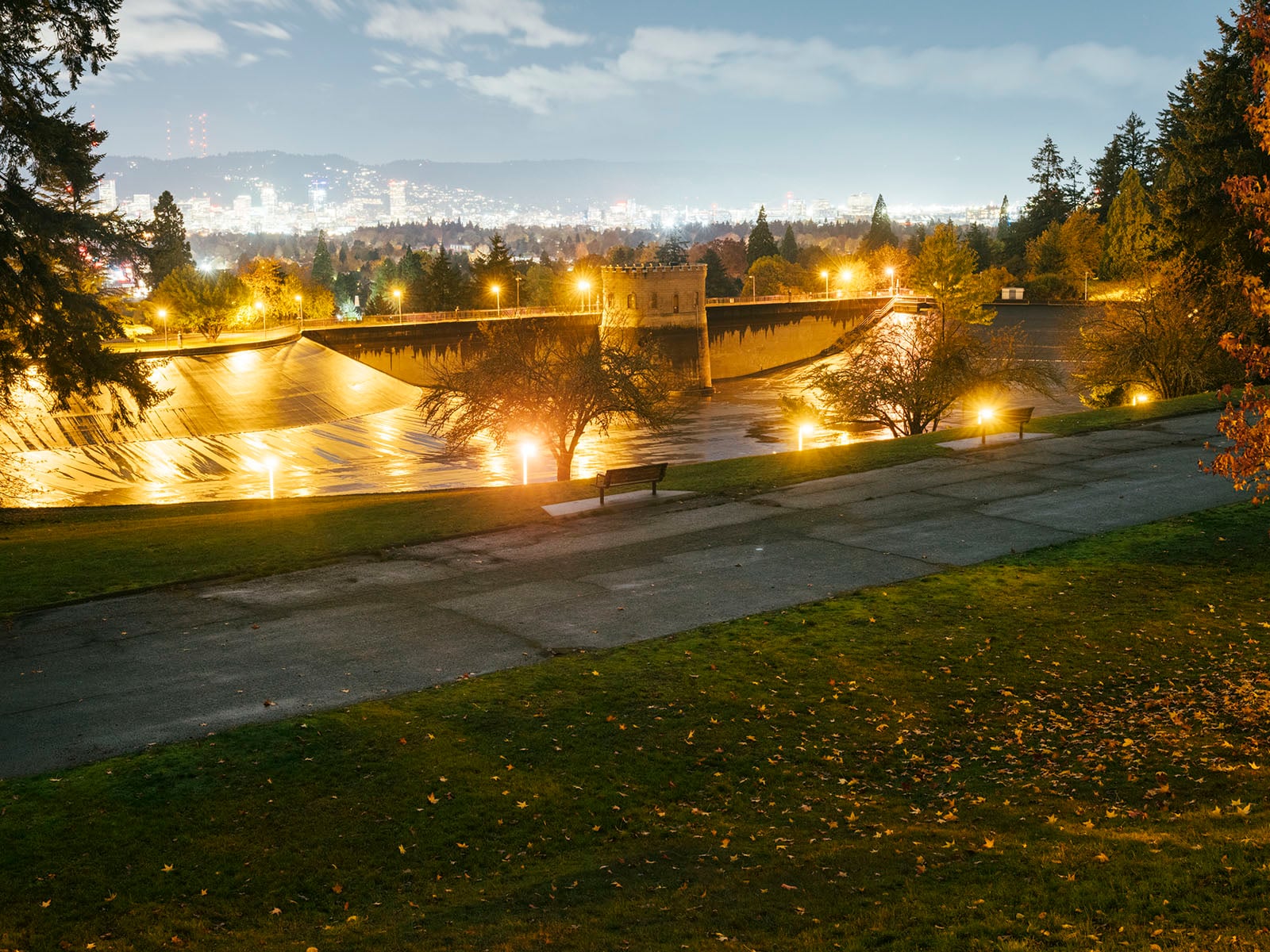 A nighttime view of a large, illuminated reservoir surrounded by trees and grassy areas, with city lights glowing in the background under a partly cloudy sky.