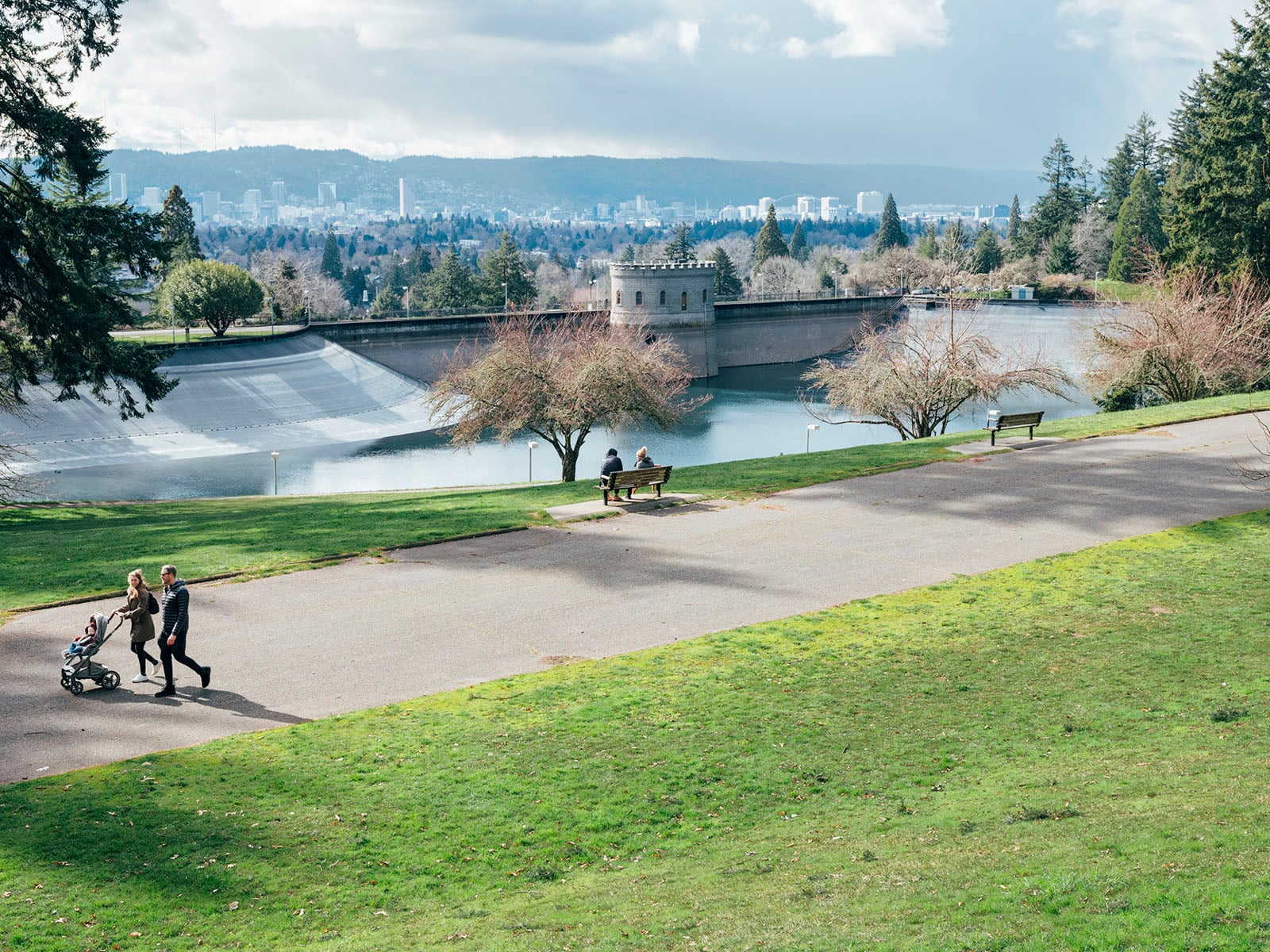 A couple pushes a stroller along a paved path in a green park, with benches and leafless trees nearby. In the background, there is a large reservoir and a city skyline under a partly cloudy sky.
