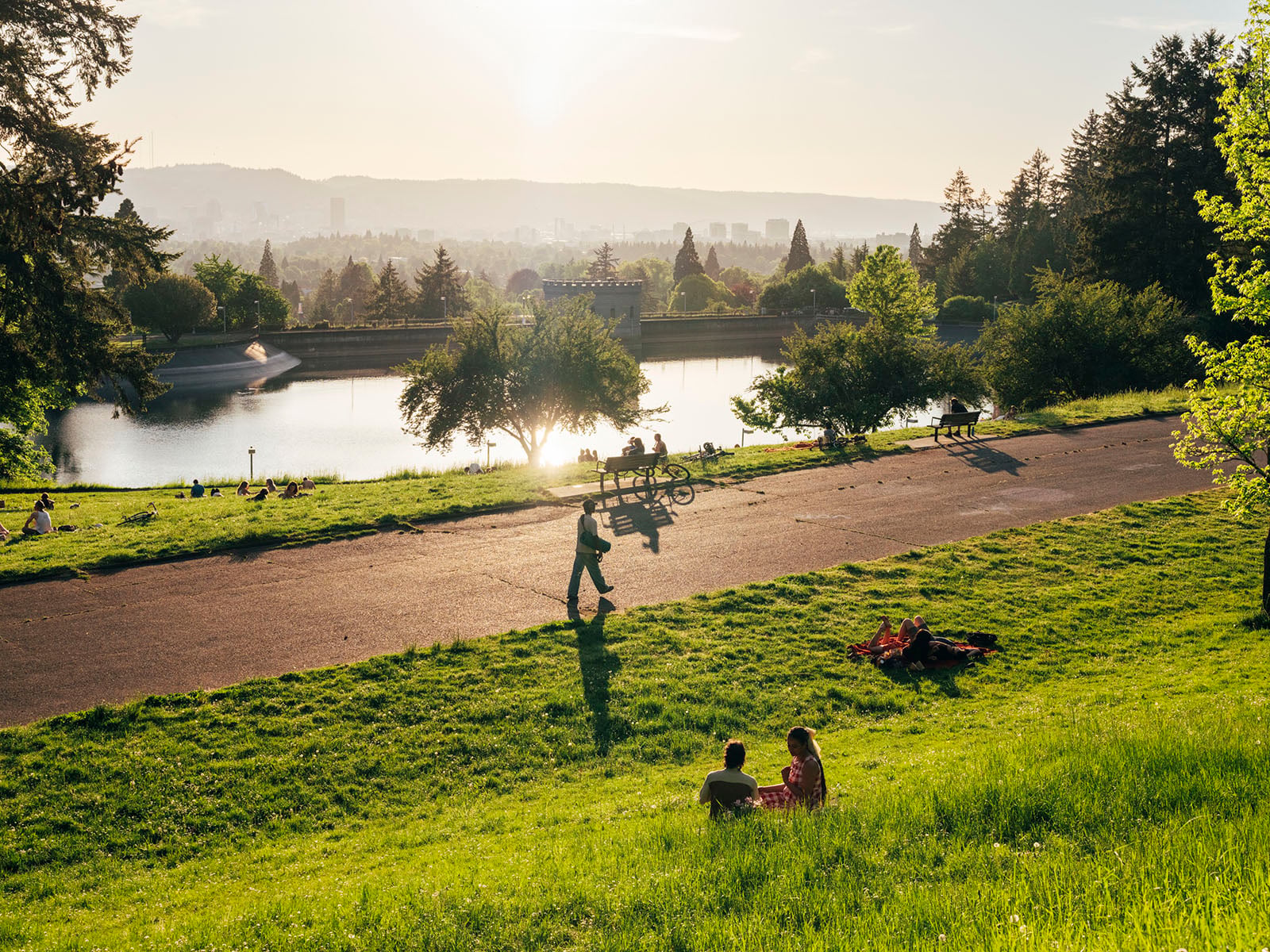 People relax on a grassy hill overlooking a reservoir at sunset, with trees, a walking path, and a city skyline in the background. Some individuals sit in groups, while others stroll or bike along the path.