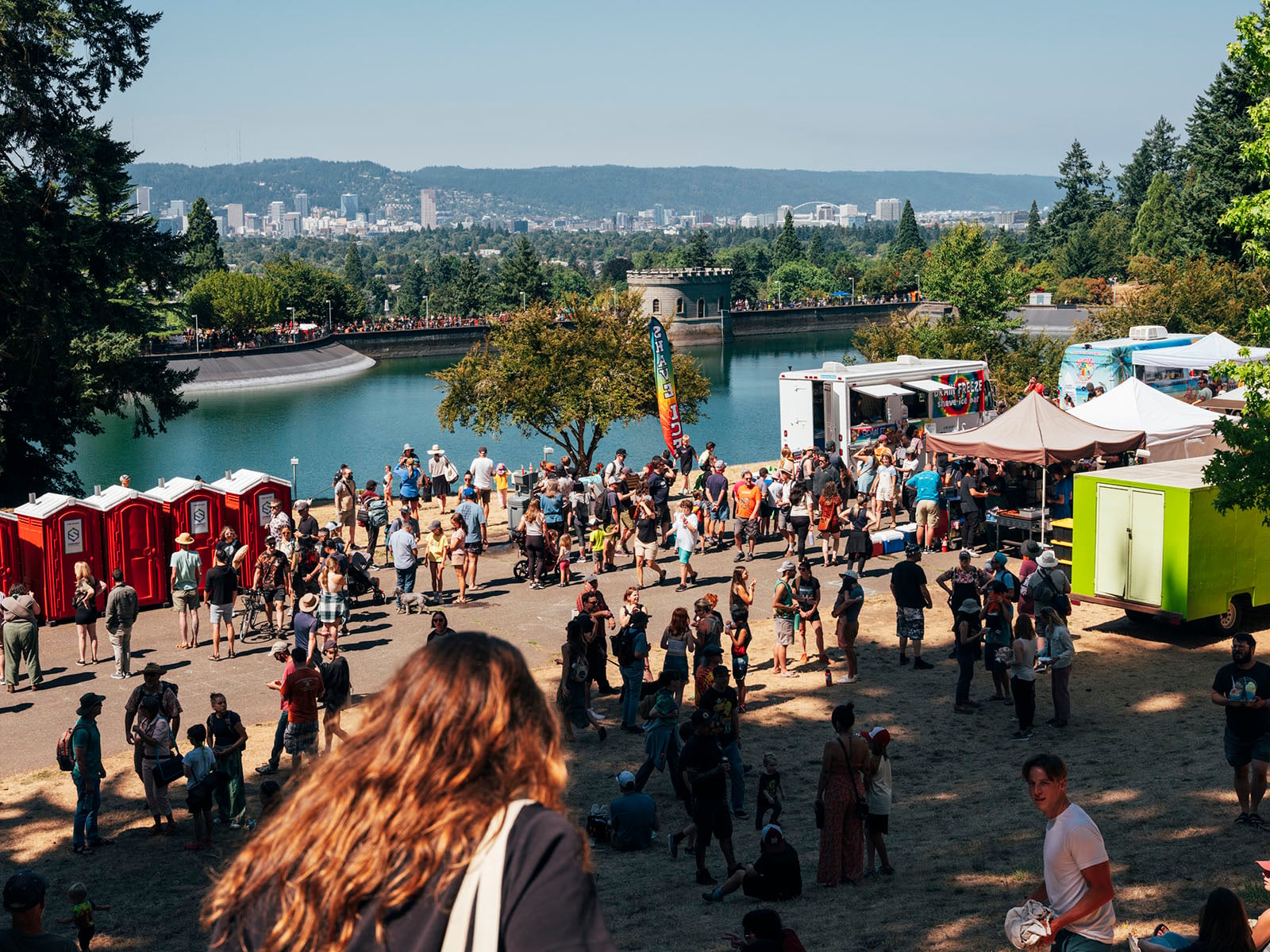 A large crowd gathers at an outdoor festival near a reservoir with food trucks, tents, and portable toilets; the city skyline and hills are visible in the background under a clear sky.