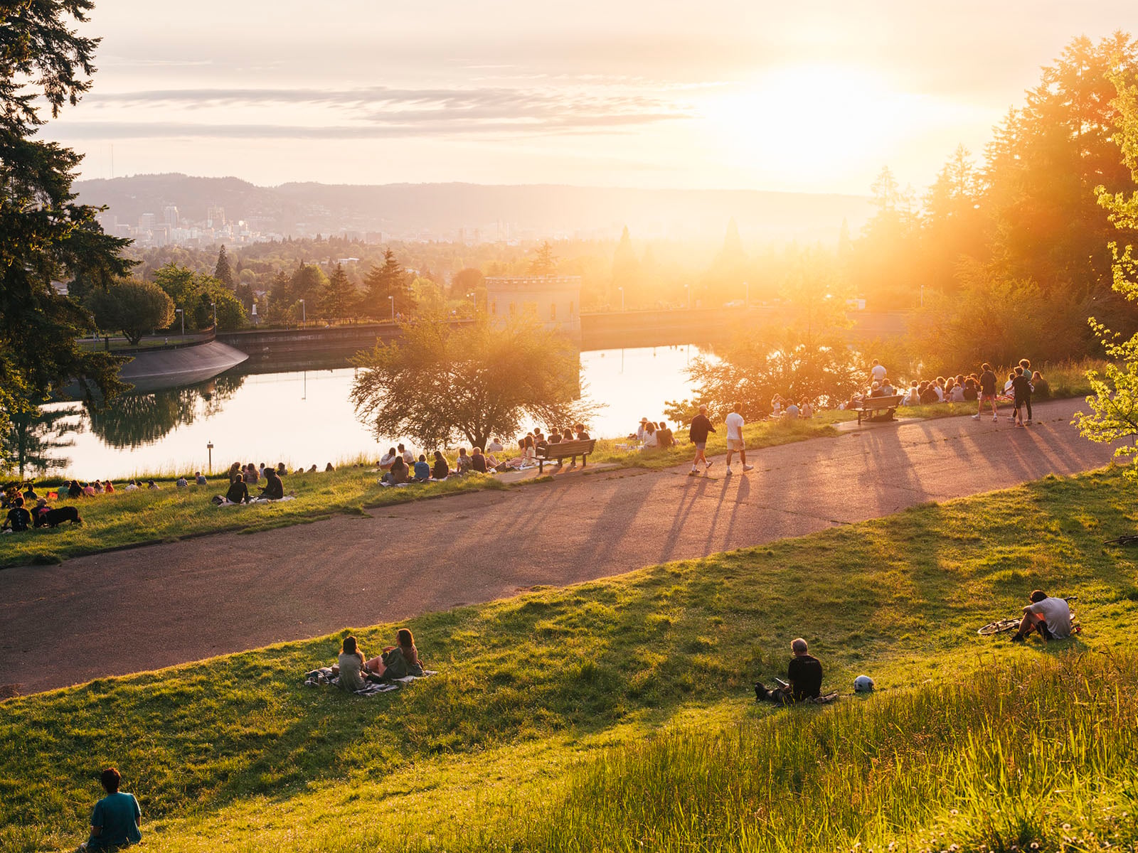 People relax on a grassy hill at sunset near a reflective reservoir, with trees, a path, and a cityscape in the background, all bathed in warm, golden light.