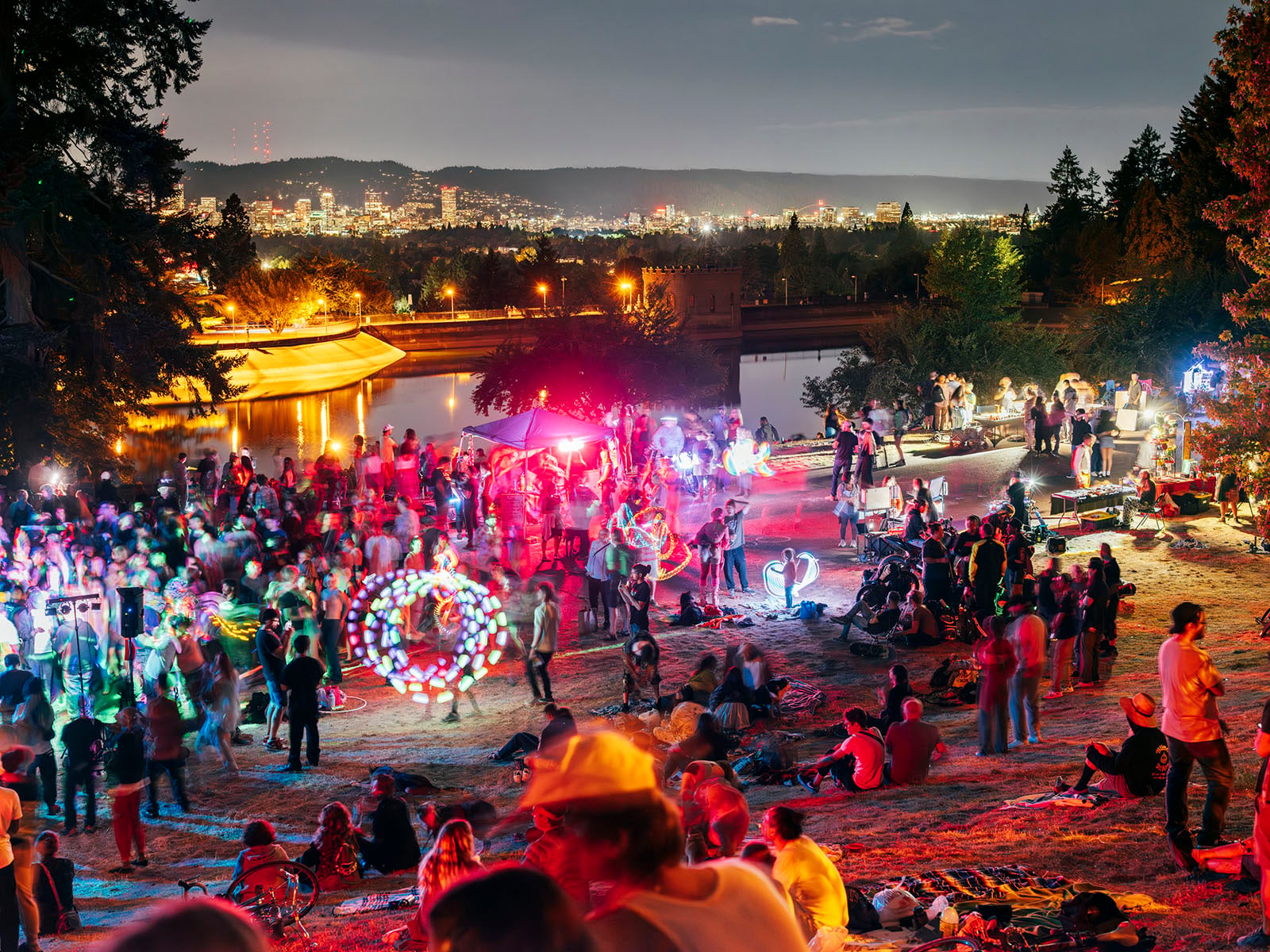 A large crowd gathers at night near a river, illuminated by colorful lights and glowing decorations. People are dancing, socializing, and relaxing on a grassy slope, with a lit cityscape visible in the background.