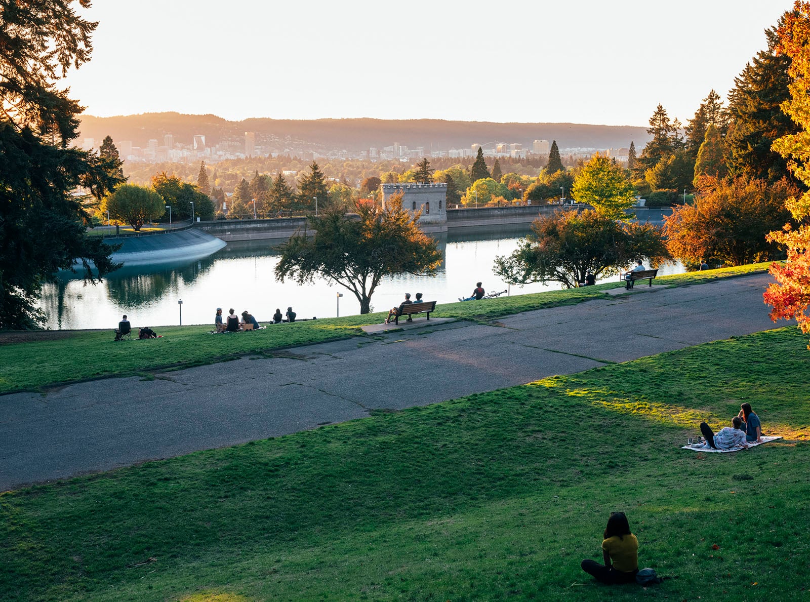 People relax on a grassy hill overlooking a large reservoir, with trees and a city skyline in the background under a setting sun. Some people sit on blankets or benches, enjoying the peaceful outdoor scene.